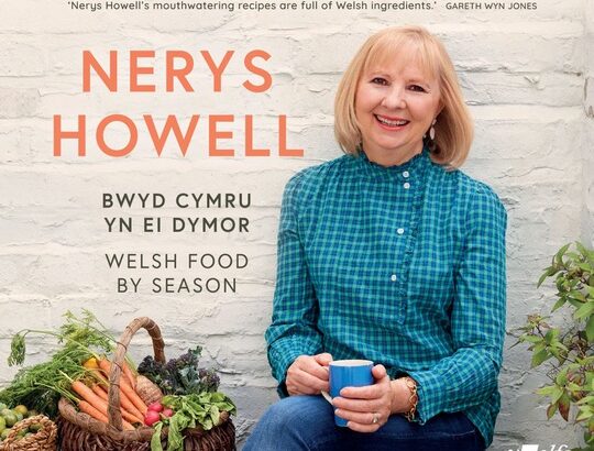 Nerys Howell (older woman in green top holding a mug) on her book cover sat next to a basket of vegetables